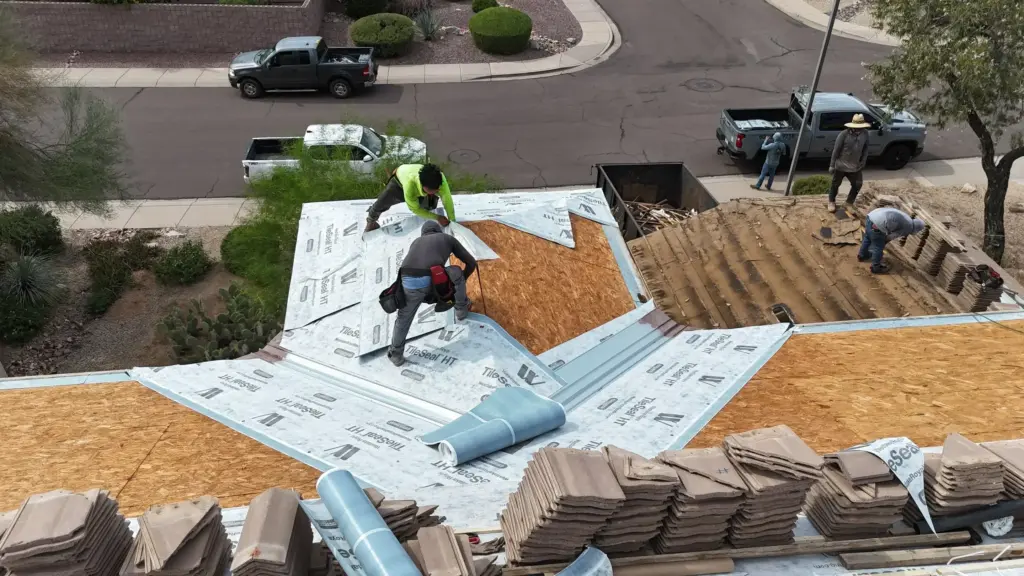 Construction workers install roof underlayment and start laying tiles on a sloped house roof from a high vantage point overlookng a suburban street.