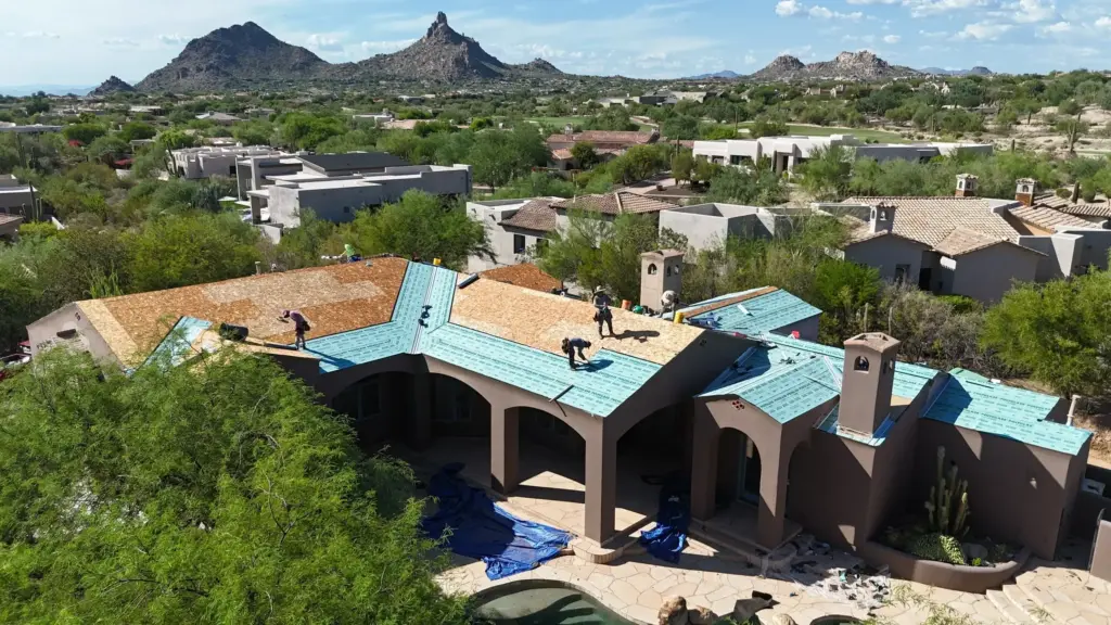 Aerial view of a desert suburb with mountains in the distance; workers install blue roof underlayment on a large house.