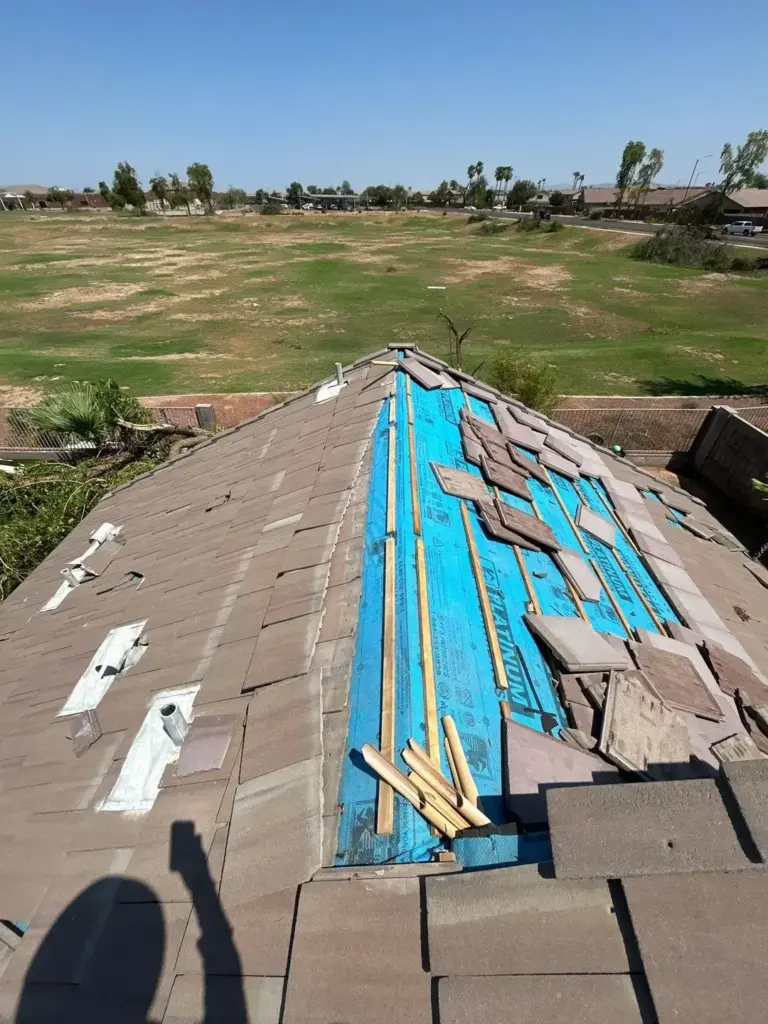 Roof under renovation with blue underlayment and scattered tiles, overlooking a grassy field and suburban houses in the distance
